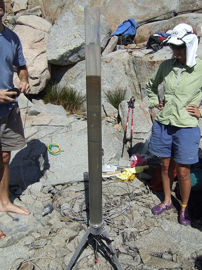 Two scientists stand on rocky shore of lake examining a lake sediment core that is in a tall transparent tube held in a metal stand.