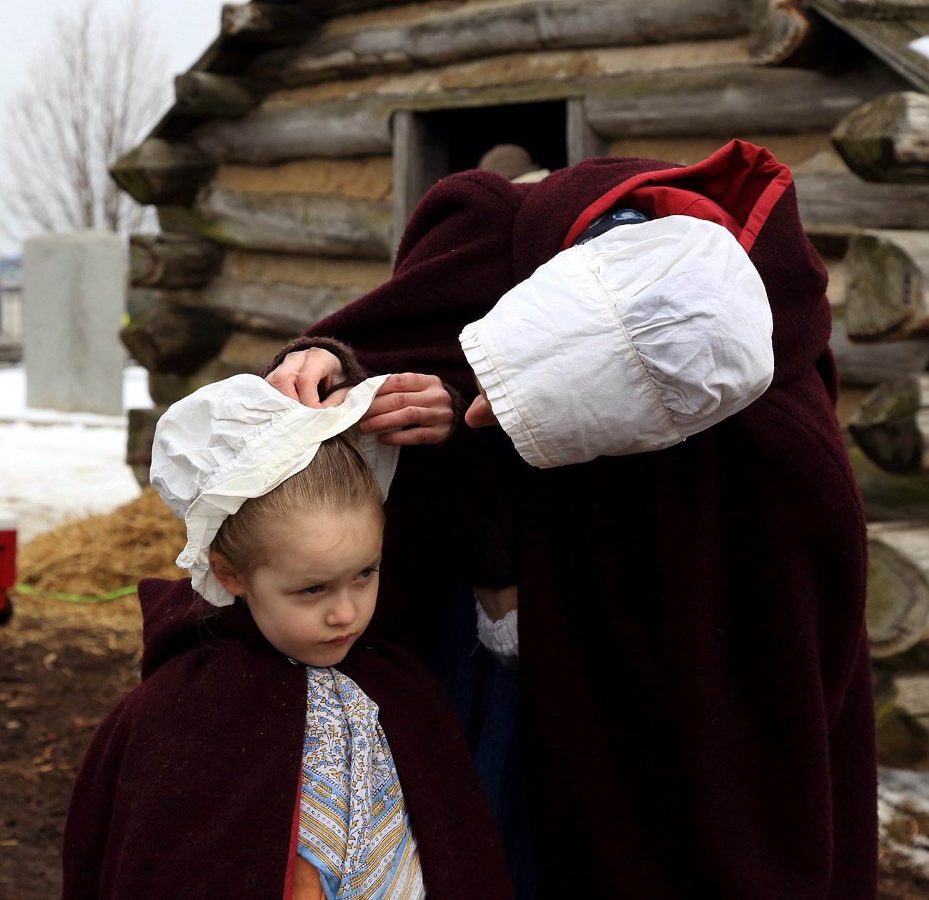 two people dressed in 18 century clothing. The parent adjusts the child's bonnet.