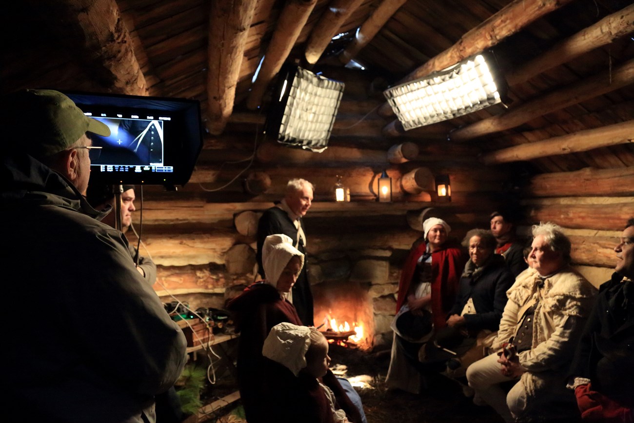 A group of people dressed in 18 century clothing sit in a small log cabin room, a modern day film crew surrounds them.