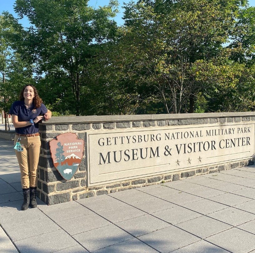 A person in a polo uniform shirt stands next to a sign that reads, "Gettysburg National Military Park Museum and Visitor Center"