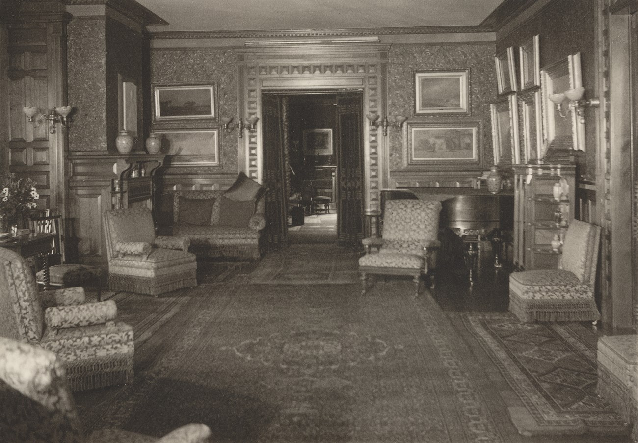 Mansion Parlor 1935 Photograph of Parlor in the Mansion, 1935. The perspective is a eye level looking down the room towards the door into the Hall. On the Left are several chairs, a sofa and the fireplace. The walls are hung with many framed paintings.