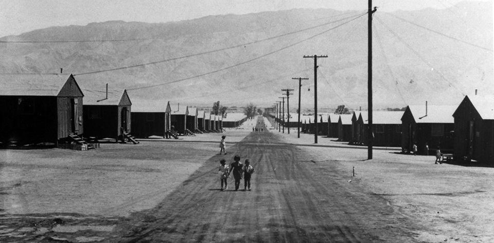 black and white photo of children walking away from us on a dusty lane between matching rows of low-slung buildings with mountains in the distance