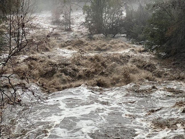 View looking downstream at muddy, fast-moving, high flow of a river swollen from large amounts of rain.