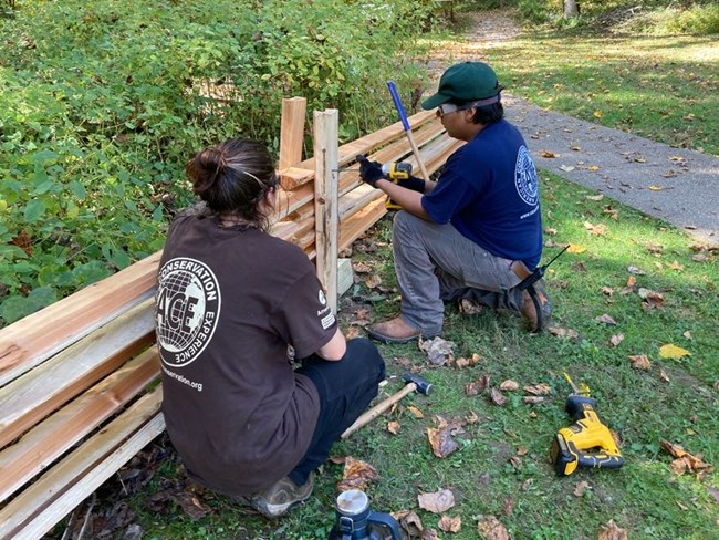 Two young individuals with tools replace a wooden fence