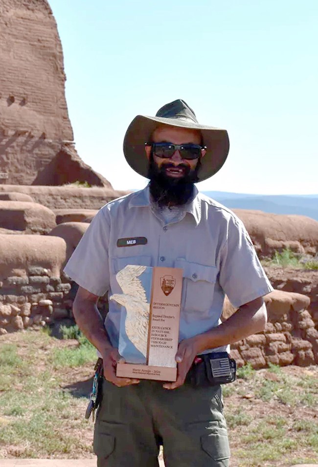 A person in a gray, collared uniform shirt, wide brimmed hat and sunglasses stands in front of a succession of short degrading mud brick walls in front of a mud brick building.