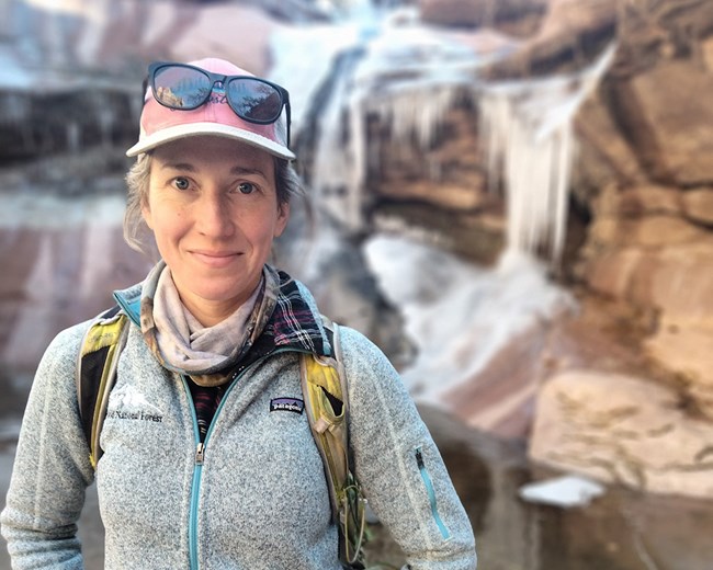 Woman wearing warm jacket, pack, and ball cap smiles as she stands in icicles of frozen water flowing over desert rocks.
