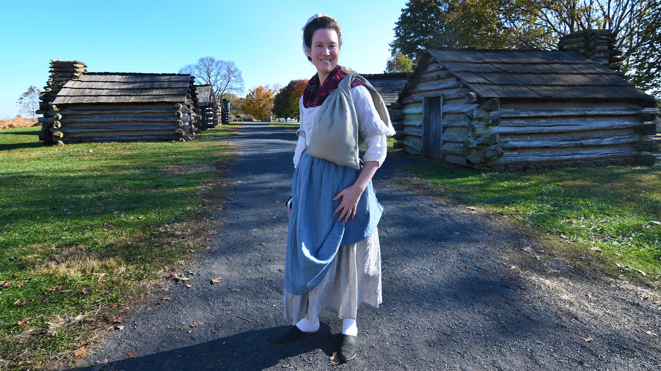 Market Wallet photograph, outdoors, smiling woman, 18th century clothing, market wallet, log huts