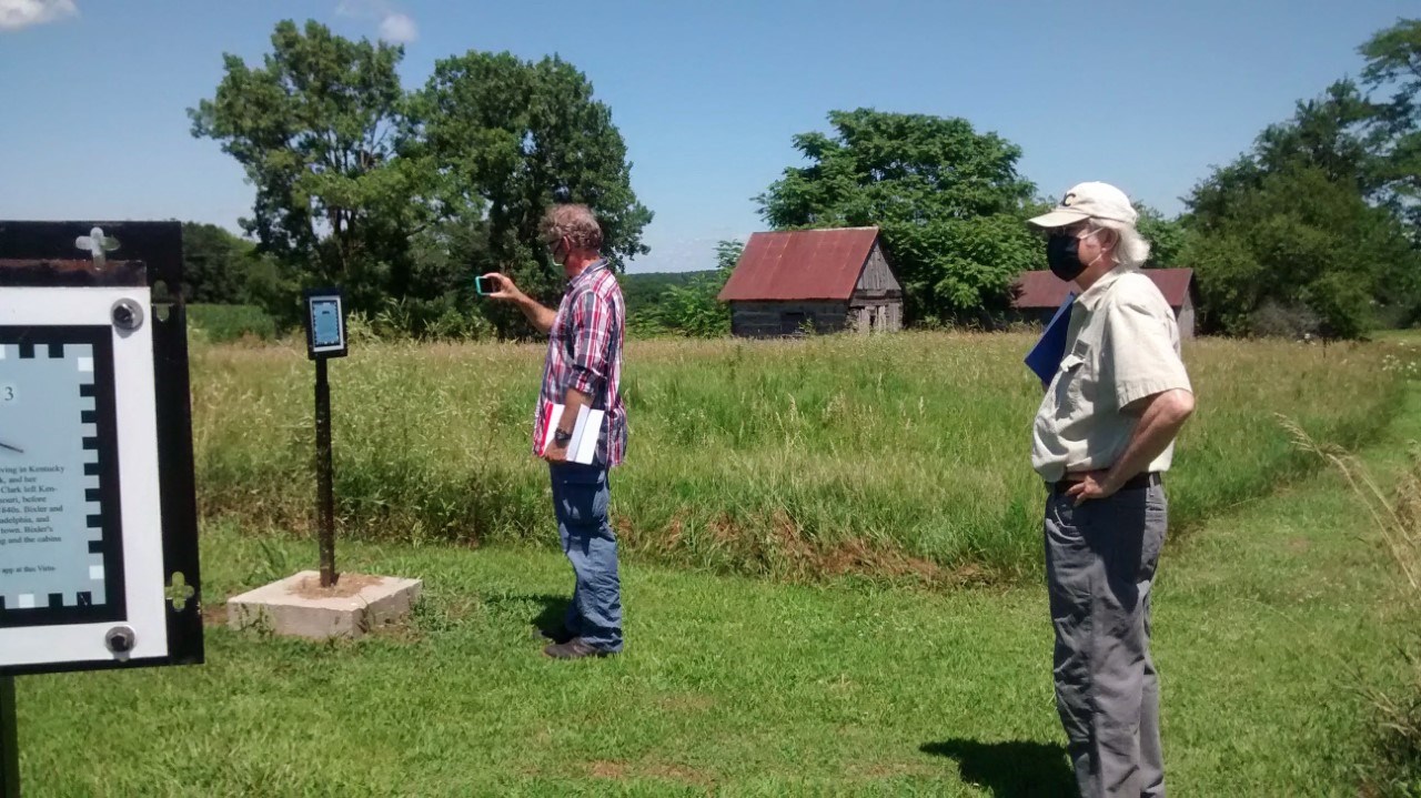 Photograph showing two people in a field with historic structures standing in tall grass in the background. One individual has his phone held up as if he is scanning a sign.