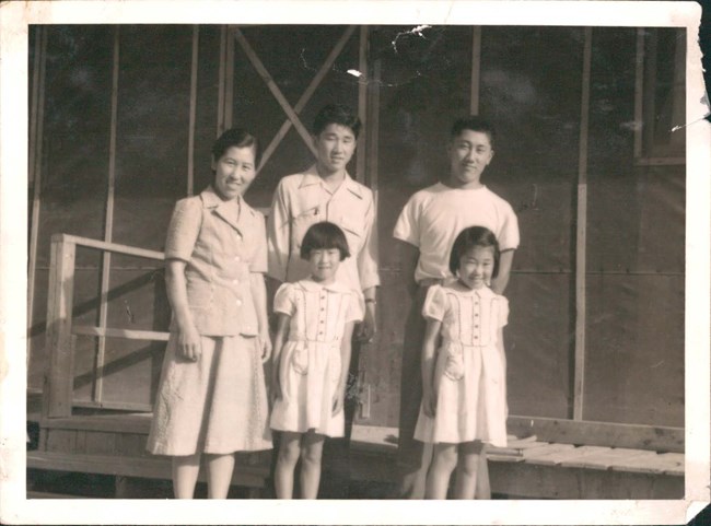 Masako, Saylo, and Tad reunited with Kazi and Aki in Poston. Courtesy of Janice Munemitsu and Chapman University, Frank Mt. Pleasant Library of Special Collections and Archives. Black and white photo of a Japanese American family: a mother, her two adult sons, and young twin girls. They pose in front of a dark wooden building.
