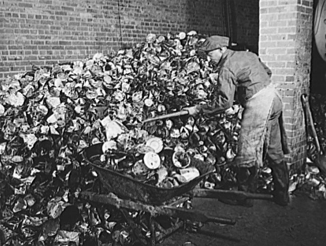 Black and white image of a white-appearing man moving a large pile of cans with a wheelbarrow and shovel.