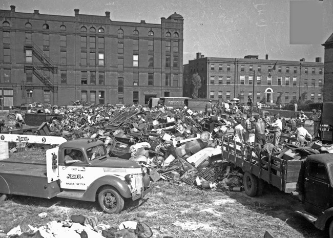 Black and white photo of a huge pile of scrap metal behind buildings in Evansville. The people in the photo look very small, indicating the size.