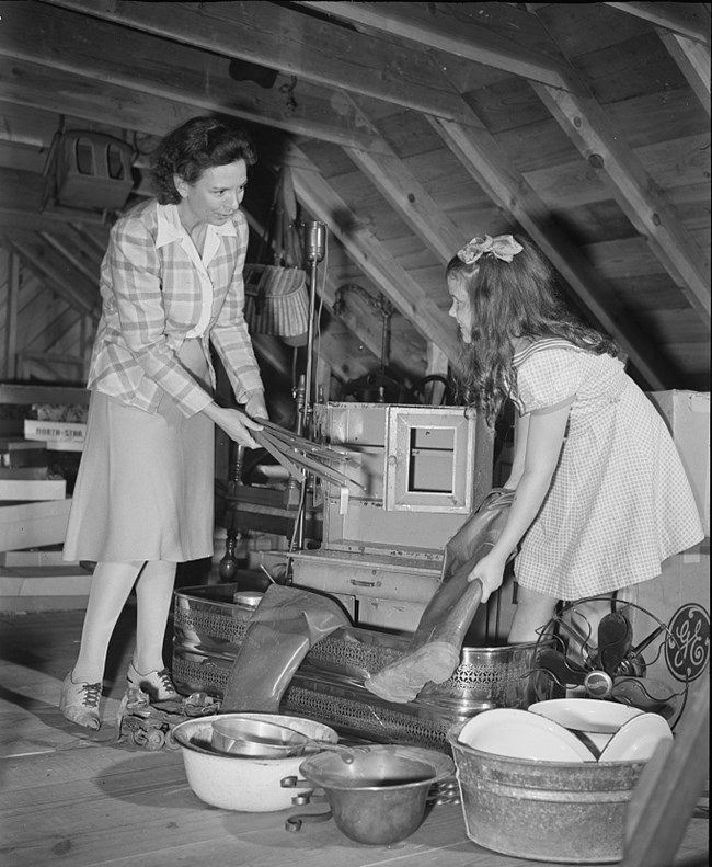 Black and white photo of two white girls in an attic. They are collecting metal items like buckets, roller skates, and fans as well as rubber boots.