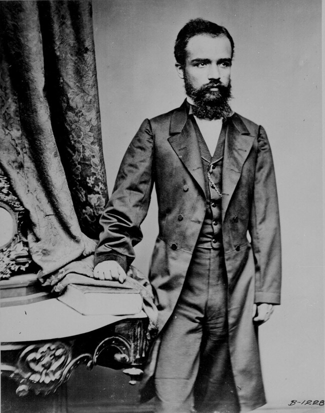 Bearded man wearing a suit and standing next to a table with a book while posing for a photo.