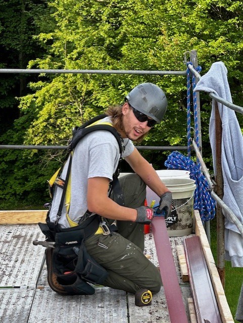 Matt Wooten marks wood with pencil and ruler at a project site. Worker wears dark sunglasses and hardhat kneeling on scaffolding as he marks a plank of wood with a ruler and pencil and looks at camera.