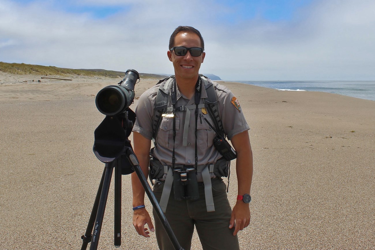Matt wearing an NPS uniform, standing beside a spotting scope on a wide, sandy beach.