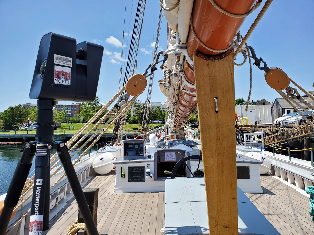 360-degree camera on deck of the Ernestina-Morrissey Boxy camera with a domed lens sits on a tripod on the deck of a schooner, just below a boom, facing the wheel and the stern.