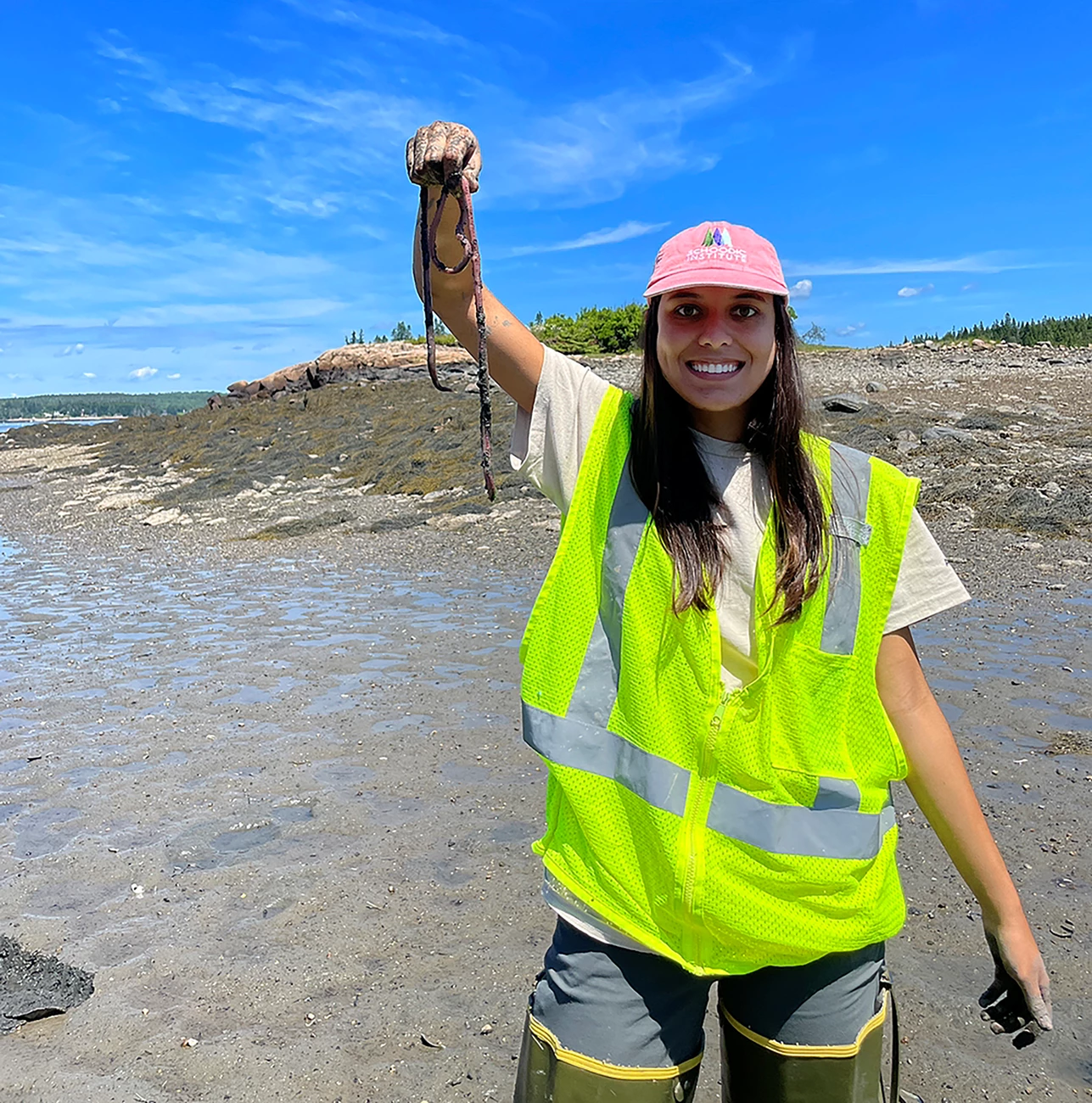 Maya Chari with marine worm Woman with hip waders and safety vest holds up a long worm while standing on a beach with marine worm