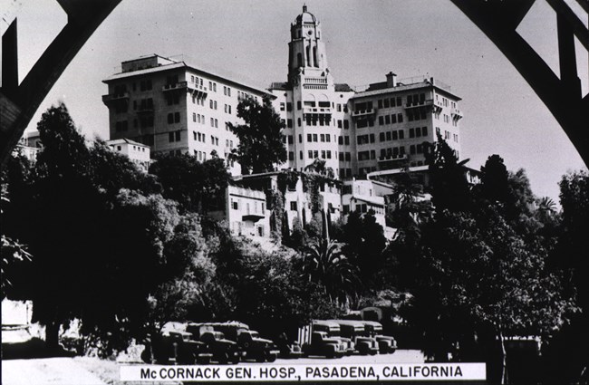 Spanish Colonial Revival style, multi-story, L-shaped building with a taller central tower. Trees and row of buses visible in the foreground.