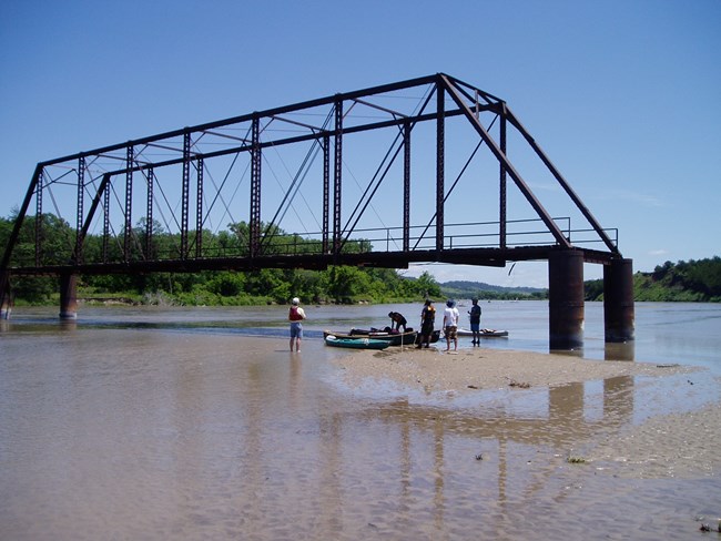 Bridge with missing span over a river, people and canoes on a sandbar looking at bridge.