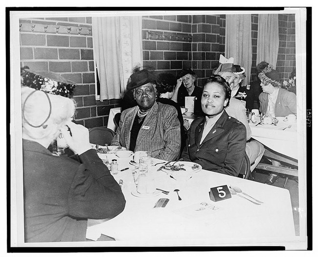 Two Black women sit next to each other at a luncheon event. The woman on the left is older, has darker skin, and is wearing a pinstriped blazer and hat with a veil. The woman on the right is younger and wears a military dress uniform.