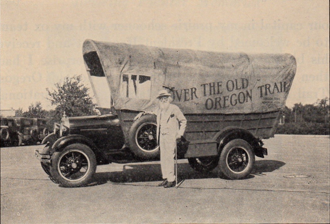 Historic photo of a man standing in front an old truck with a covered wagon on the back.