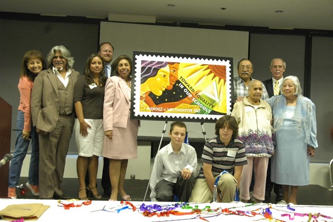 Sylvia Mendez and relatives of plaintiffs gather around an enlarged poster of the stamp displayed on an easel. The stamp features two figures in profile reading a book against a night sky. The sun shines down from another corner.