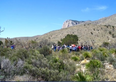 A group of tribal members gather in a landscape of green scrub brush.
