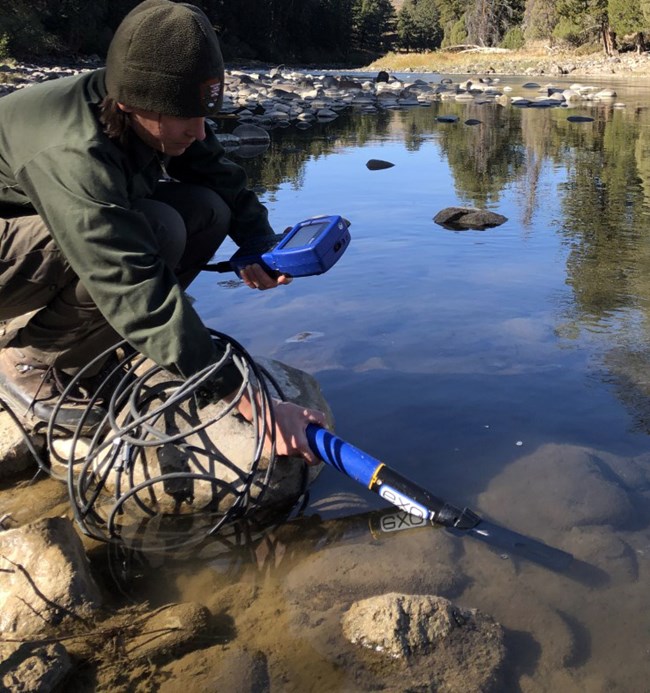 A person crouched down by flowing water holding water quality equipment.