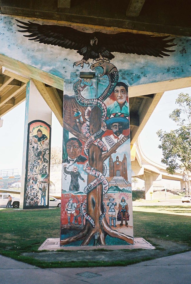 Colorful mural on a T-shaped highway pillar in Chicano Park showing an interpretation of Mexican history, including a central tree encircled by snakes and an eagle. Figures include conquistadores, missionaries and Mexican Revolutionary leaders.