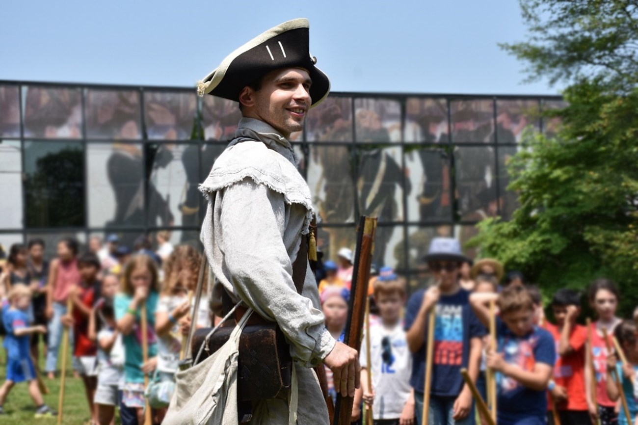 A person dressed in a Revolutionary War Uniform stands with a musket in front of a crowd.