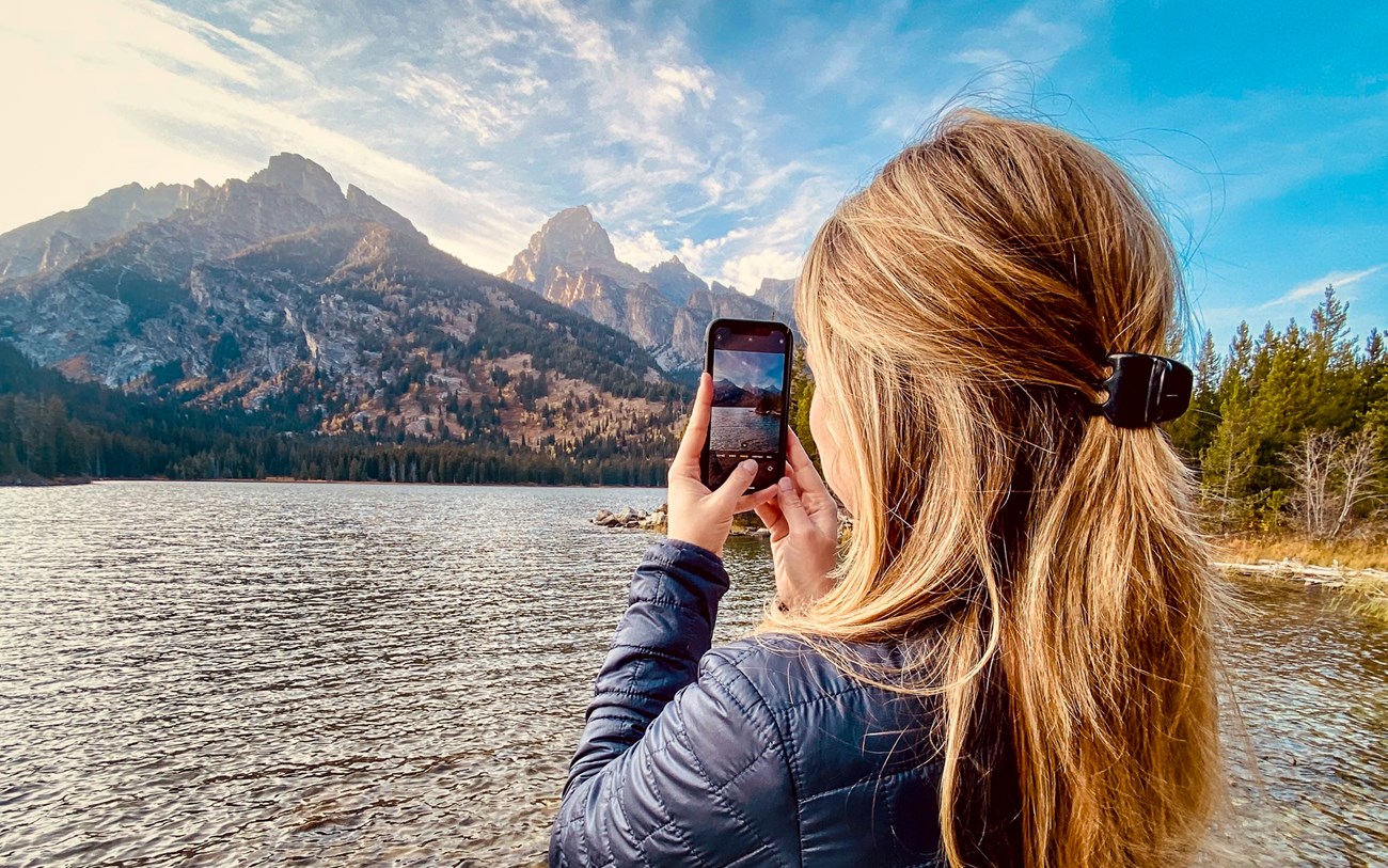 A visitor captures a photo on their phone at Taggart Lake with the Teton Mountain Range and blue skies