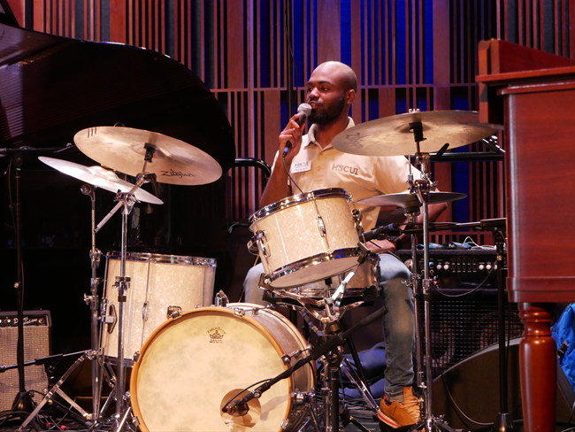 Hunter speaking to audience from behind drum set sharing his knowledge on jazz during a Down On Their Luck (a weekly orchestra of the Arrowhead Jazz Band)