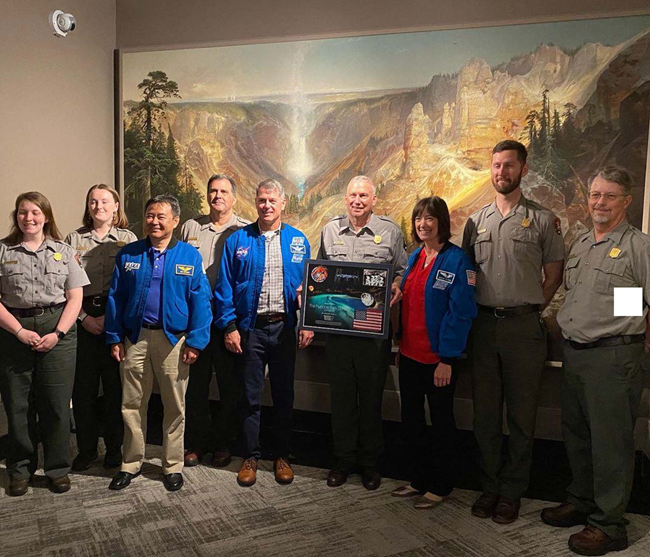 Astronauts and park rangers pose in front of Thomas Moran painting. Several astronauts and park rangers pose in front of a large colorful painting of the Grand Canyon.