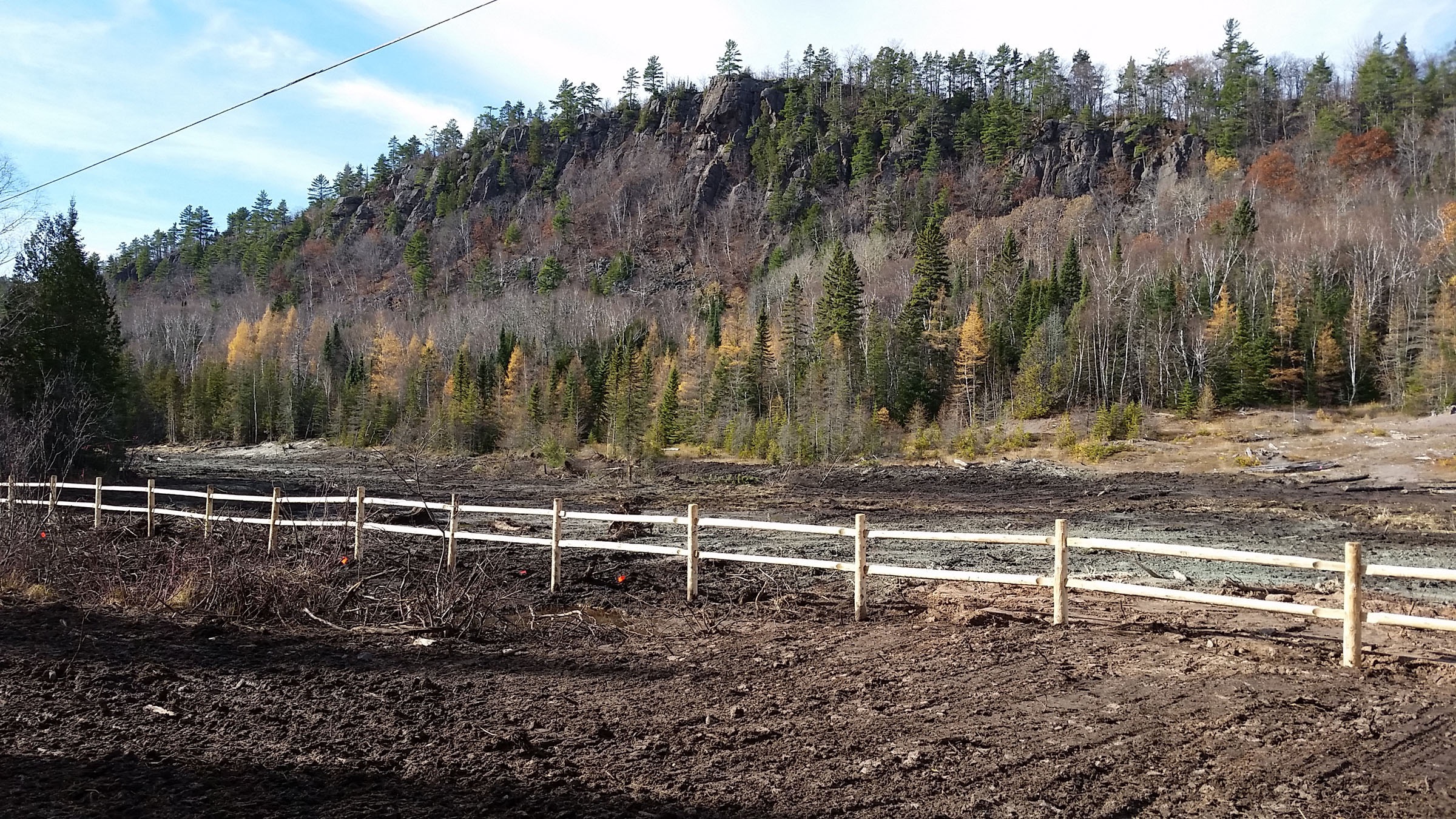 Leafless autumn image of a forested, gray rocky bluff in the background with a wooden fence running across the foreground.