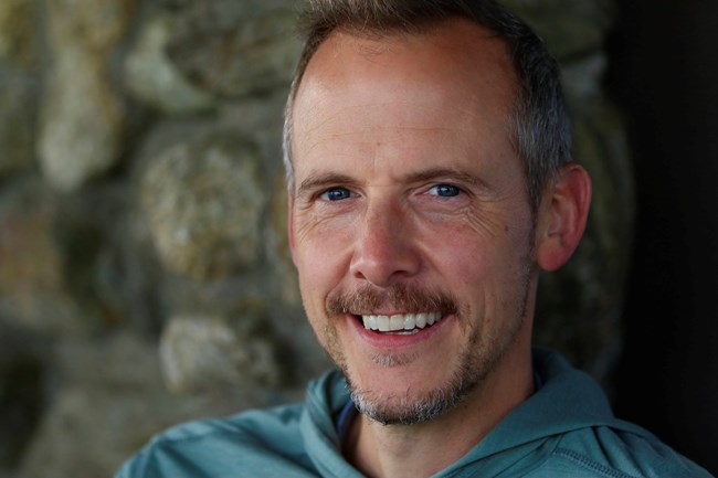 Smiling man with gray-speckled short hair and beard wearing a green hoodie in front of a stone wall