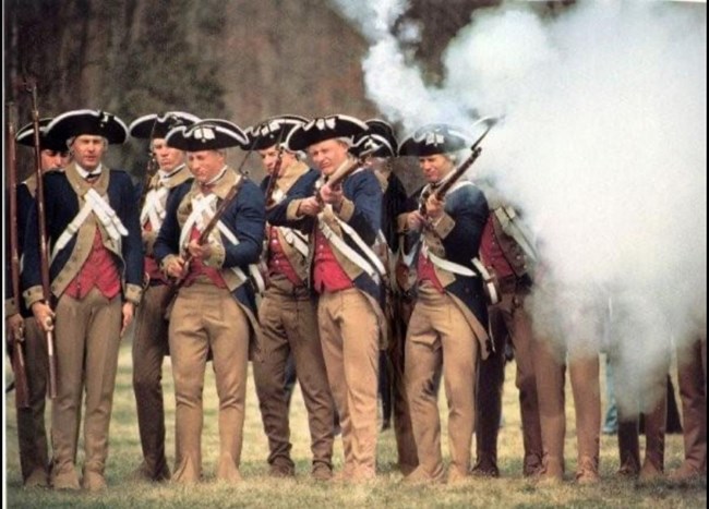 Mike Horton.3 Several men in revolutionary war outfits holding long guns. Smoke obscures the right side of the photo.