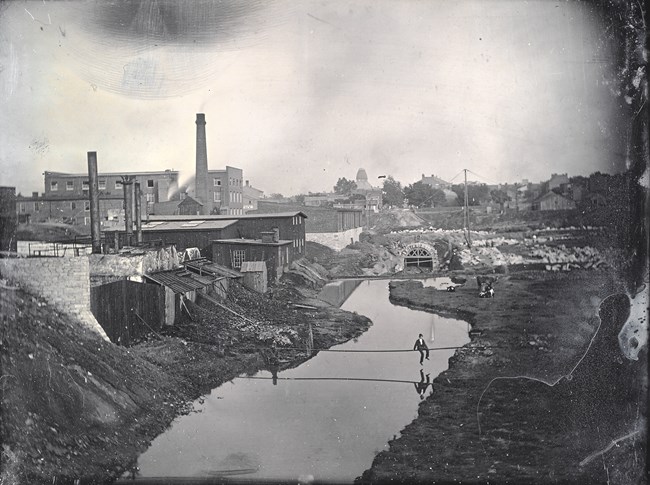 Photo of an urban area with buildings, a smoke stack, and a creek running through the middle of the photo.