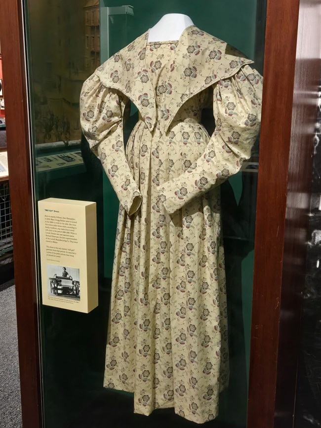 Mill Girl Dress Exhibit case holds a dress made of a yellow fabric with a floral design. The dress features puffy sleeves and a "v" shaped collar