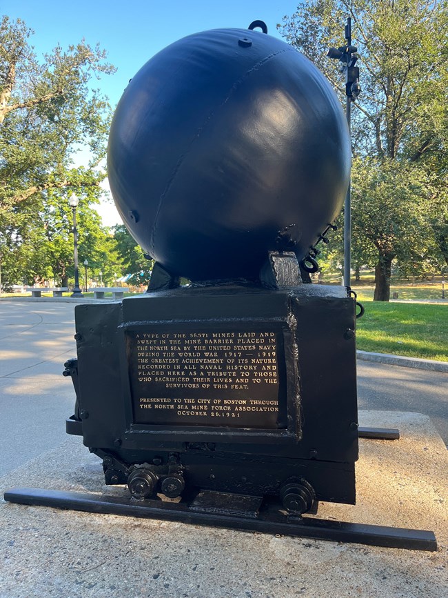 A memorial of a round ocean mine on a pedestal with an inscription below it.