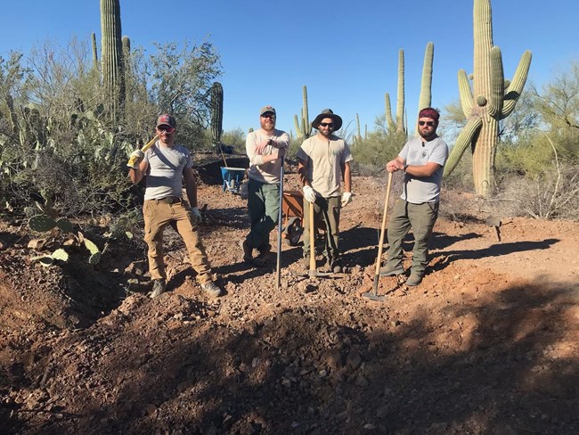 A crew of four NPS workers standing at a former 55' deep AML site. There are wheelbarrows and cacti behind them.