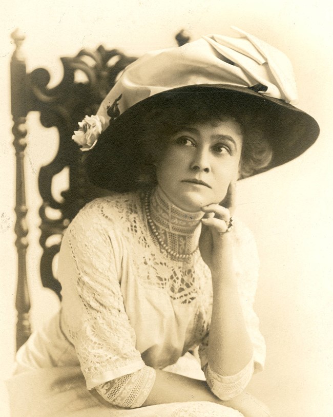 Photo of a woman, seated, with hand on chin, with broad-brimmed hat and white dress
