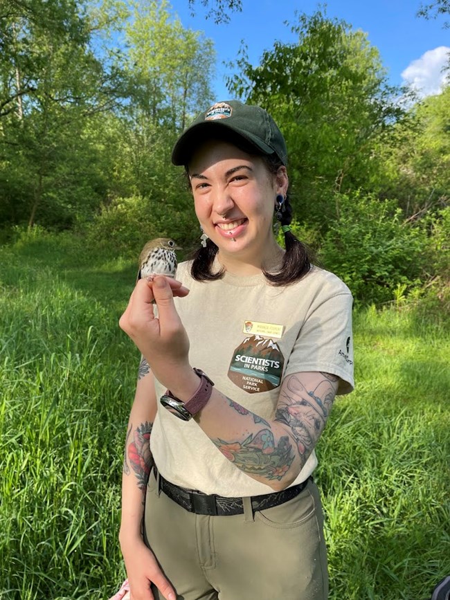 Miranda with ovenbird A woman in khaki uniform and green hat stands smiling outdoors holding a small brown and white speckled bird in her hand.