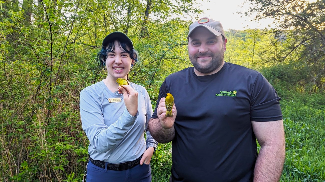 Miranda and Dan from SMP Two people stand side by side smiling outdoors, each holding a small yellow bird in their hand.