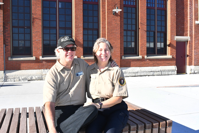 A Caucasian man and woman sit on a bench in volunteer uniforms in front of a red stone building.