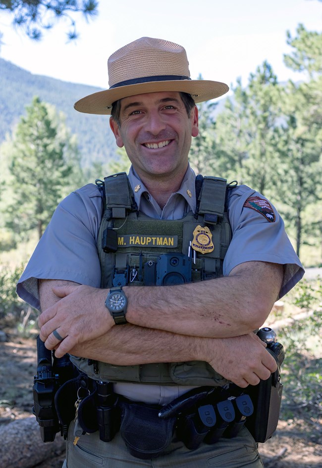 A smiling uniformed person, with a straw, flat-brimmed hat, in a forest setting.
