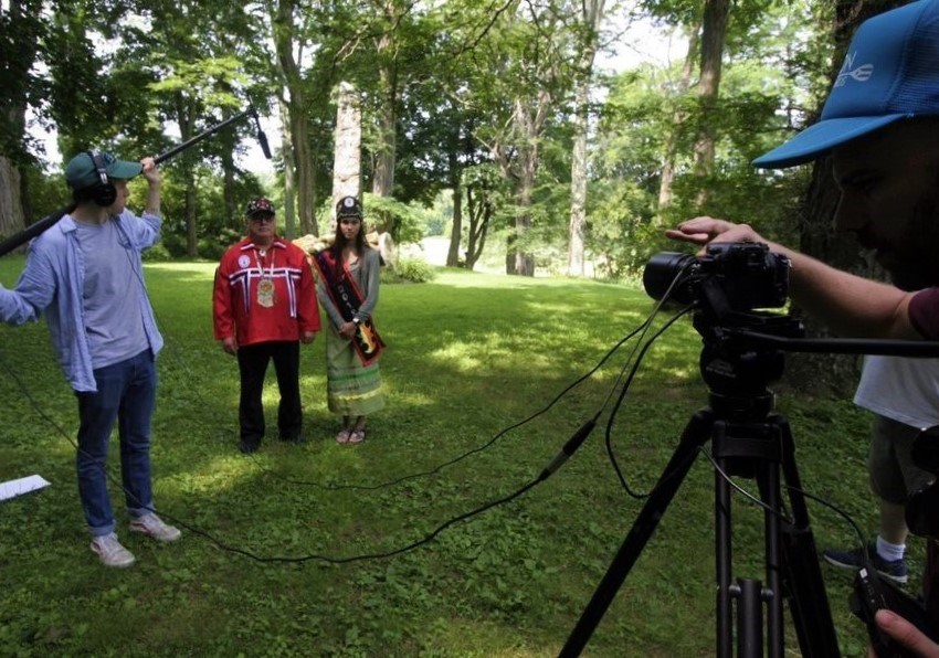 Older man and young woman stand in front of camera crew in open green space surrounded by woods. They wear traditional Mohican clothing.