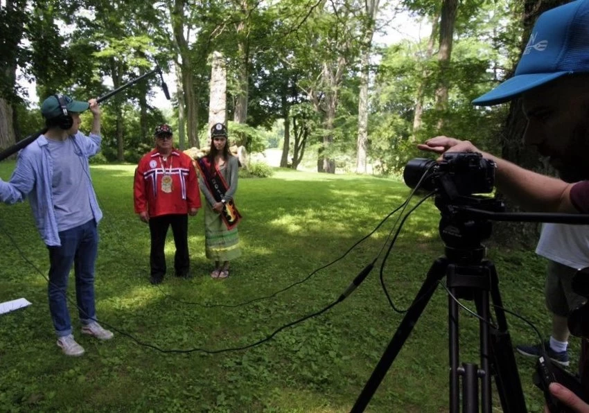Mohican Heritage Walking Tour, from Stockbridge-Munsee Community Band of Mohicans & Upper Housatonic NHA Older man and young woman stand in front of camera crew in open green space surrounded by woods. They wear traditional Mohican clothing.