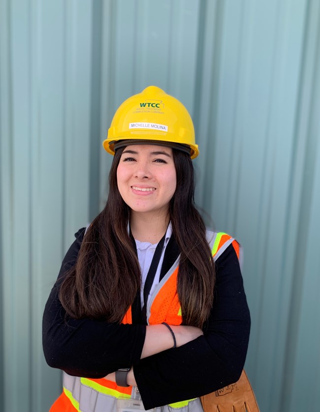 Michelle Molina A woman with long brown hair wears a yellow hardhat, orange safety vest and holds a leather glove in her hand. She stands in front of a metal building with her hands crossed in front of her.