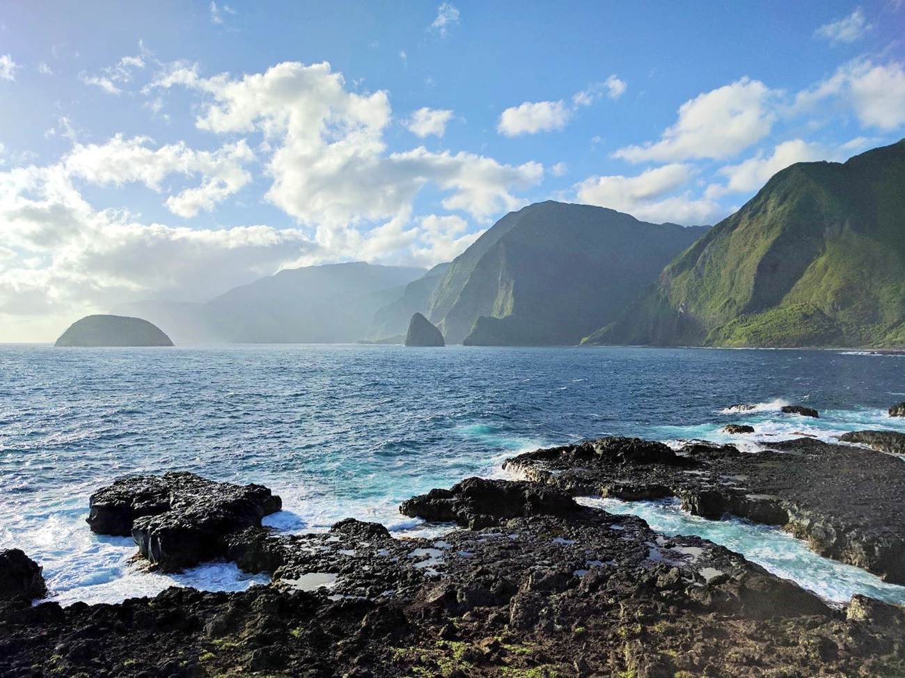 Northern coast of Molokaʻi Looking from a rocky, tide-pool-dotted shore across sparkling blue-green water at small islands and towering green sea cliffs, the furthest of which are cloaked in clouds.