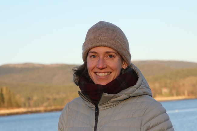 Headshot of a woman in a gray puffy jacket and beanie, in front of a hilly landscape with a lake.